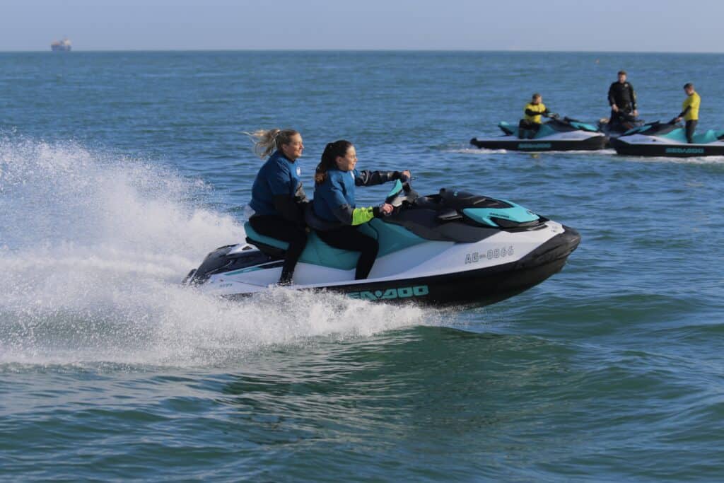 Two women riding a jet ski on the ocean with safety gear, enjoying water sports.
