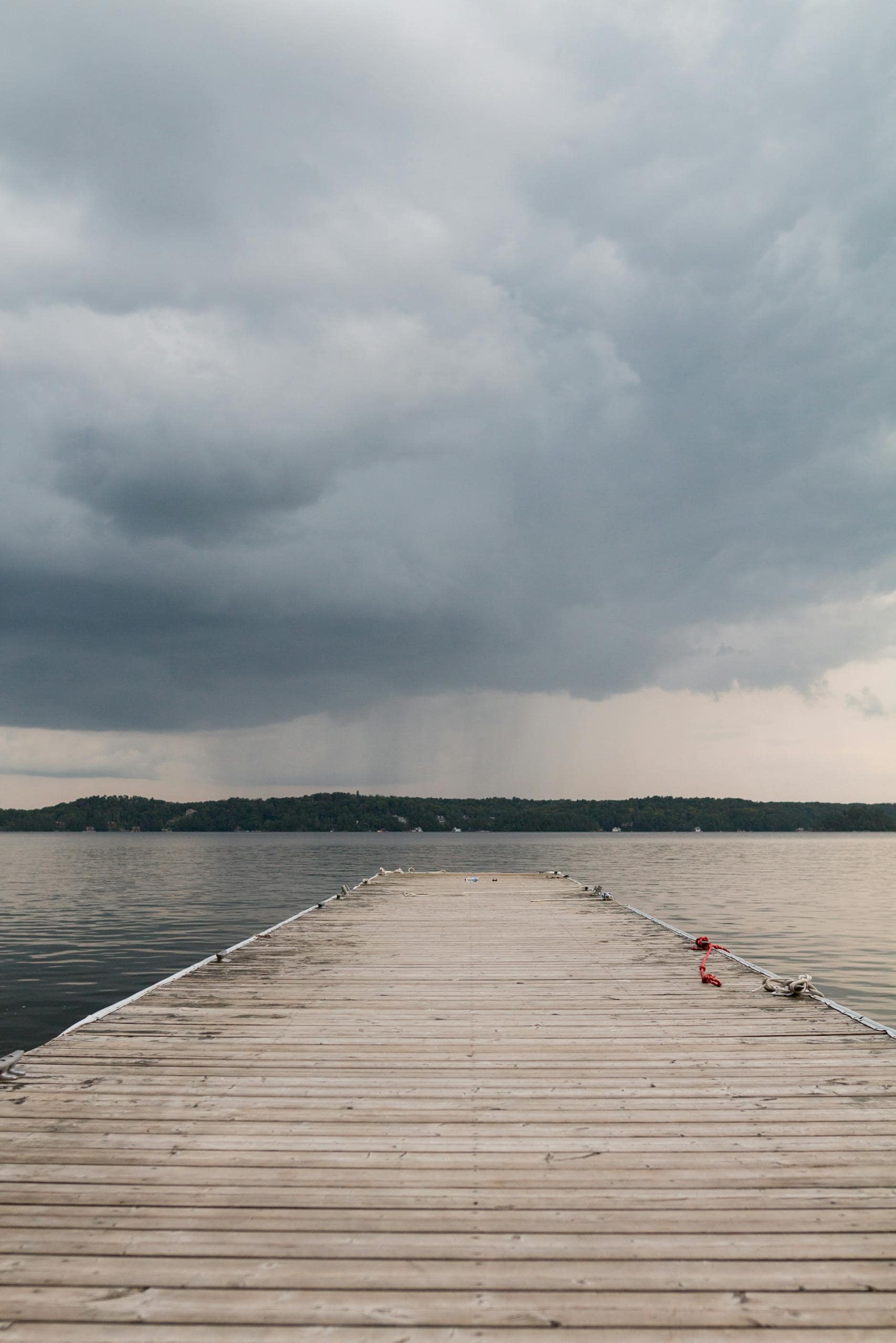 Wooden berth leading to water with deep place against hilly terrain with trees and houses under cloudy sky with coming rain